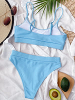 Blue bikini set on a white surface with a hat, avocado, and wooden board in the background.