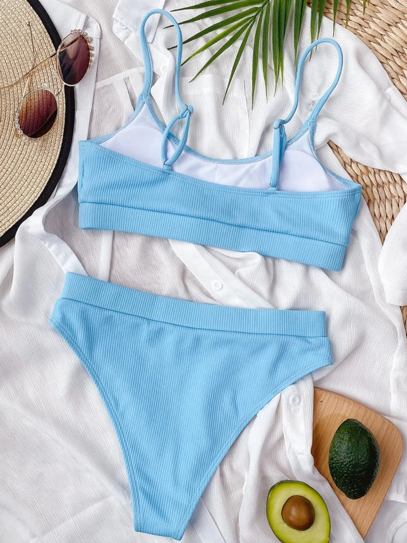 Blue bikini set on a white surface with a hat, avocado, and wooden board in the background.