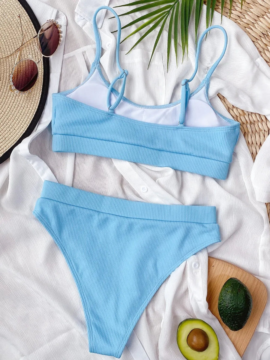 Blue bikini set on a white surface with a hat, avocado, and wooden board in the background.