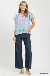 Woman wearing a light blue blouse with green floral embroidery and dark blue jeans on a white background.