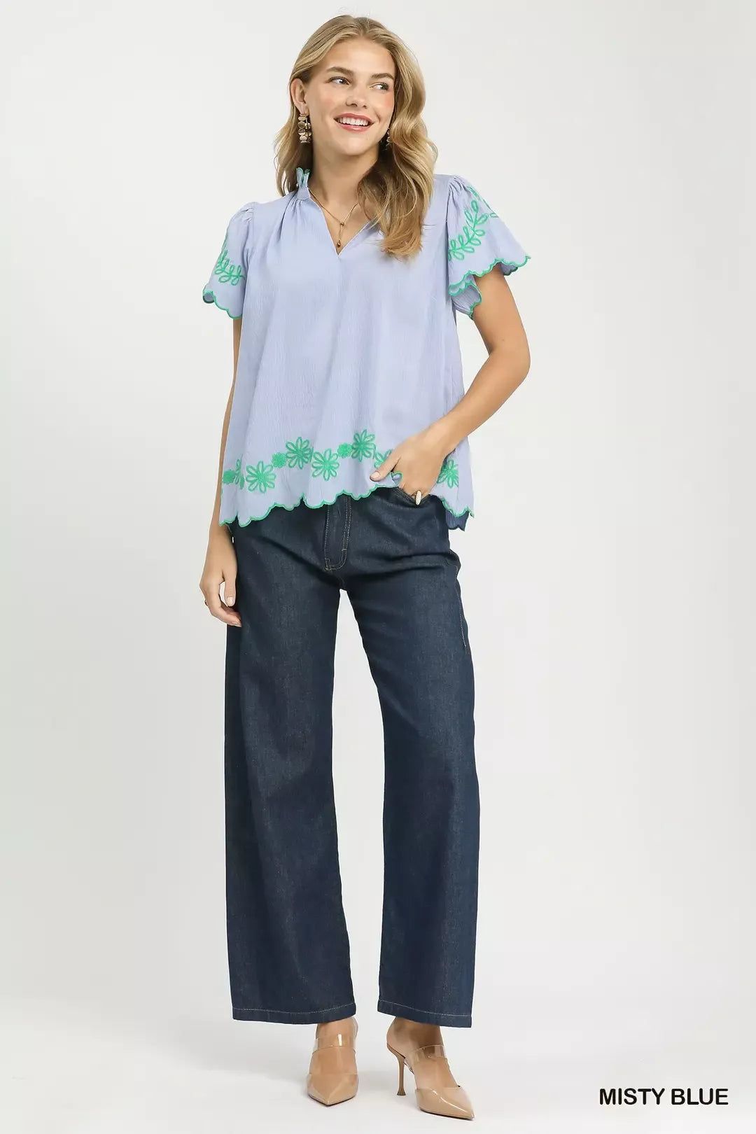 Woman wearing a light blue blouse with green floral embroidery and dark blue jeans on a white background.