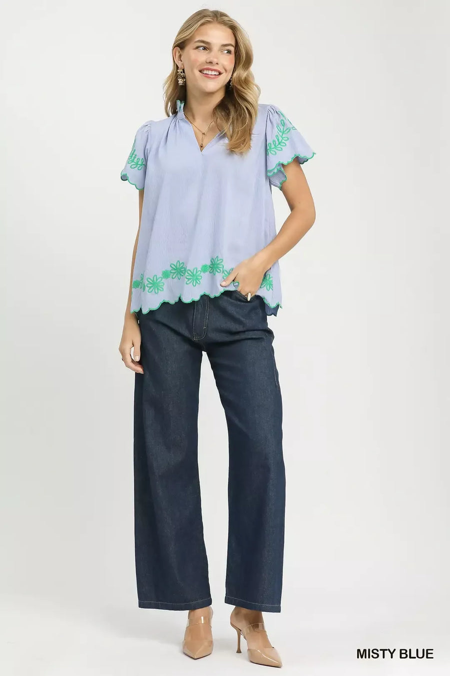Woman wearing a light blue blouse with green floral embroidery and dark blue jeans on a white background.