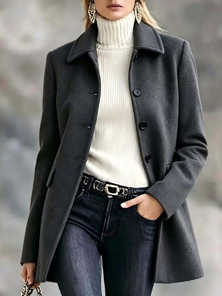 A woman wearing a dark charcoal gray wool-blend coat over a cream ribbed turtleneck sweater, paired with dark blue jeans and a black belt with a gold buckle.