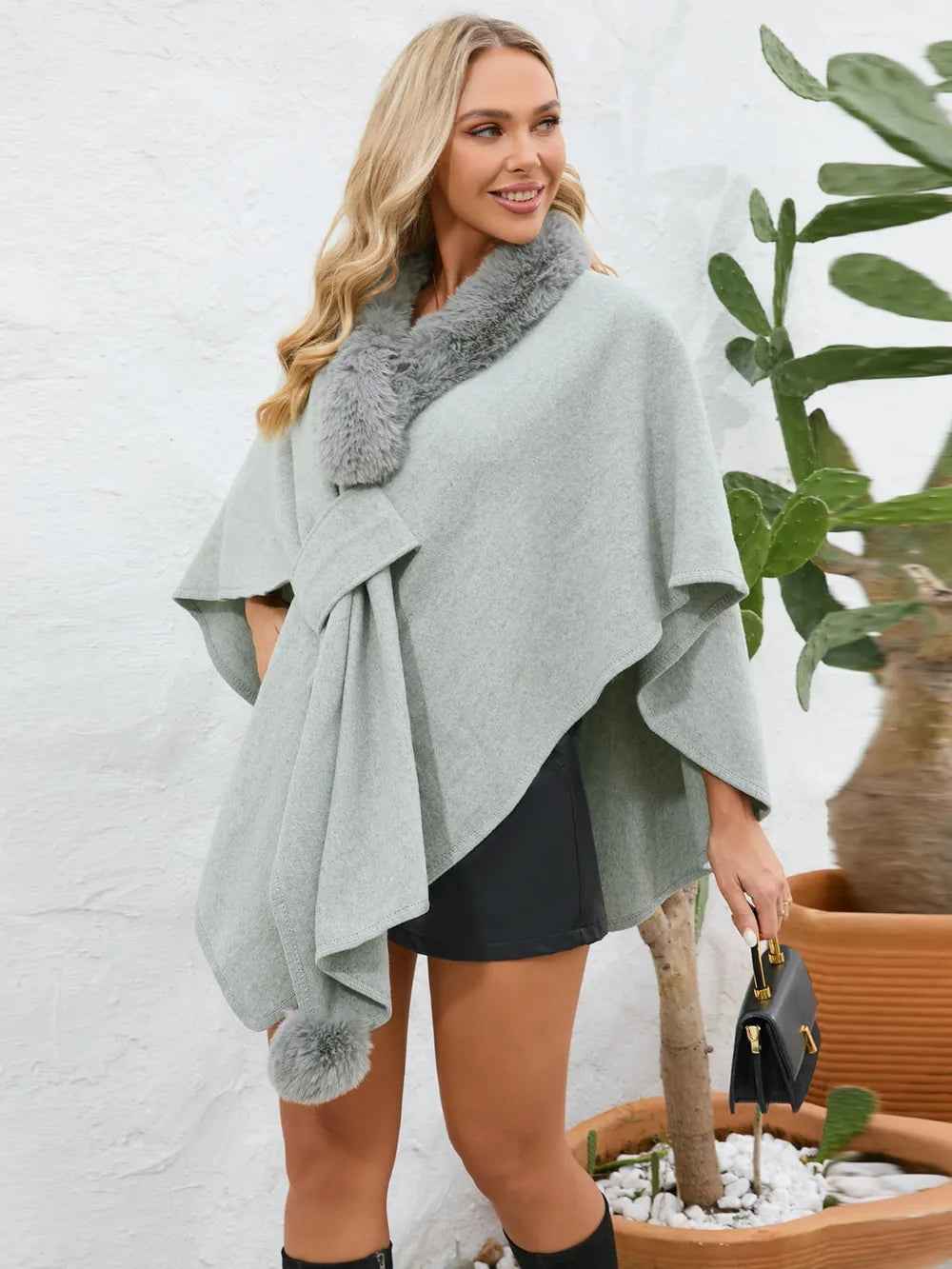 Woman wearing a gray poncho with fur trim standing next to a potted plant.
