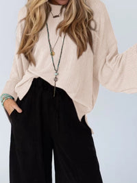 Person wearing a beige sweater and black pants with a plain background