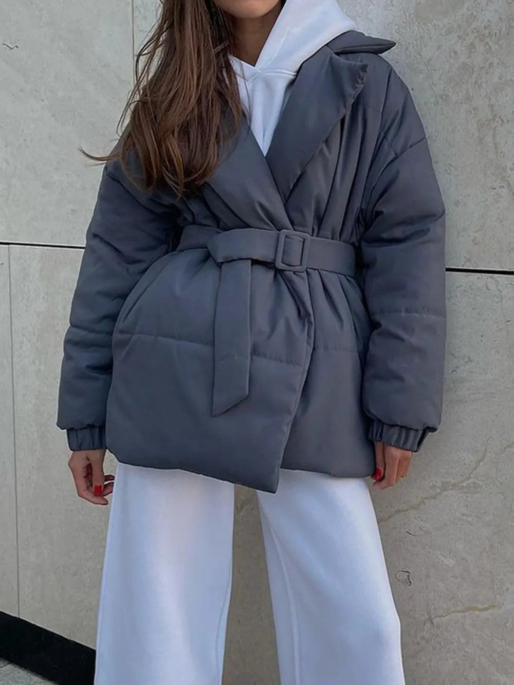 Person wearing a gray puffer coat with a belt against a neutral wall.