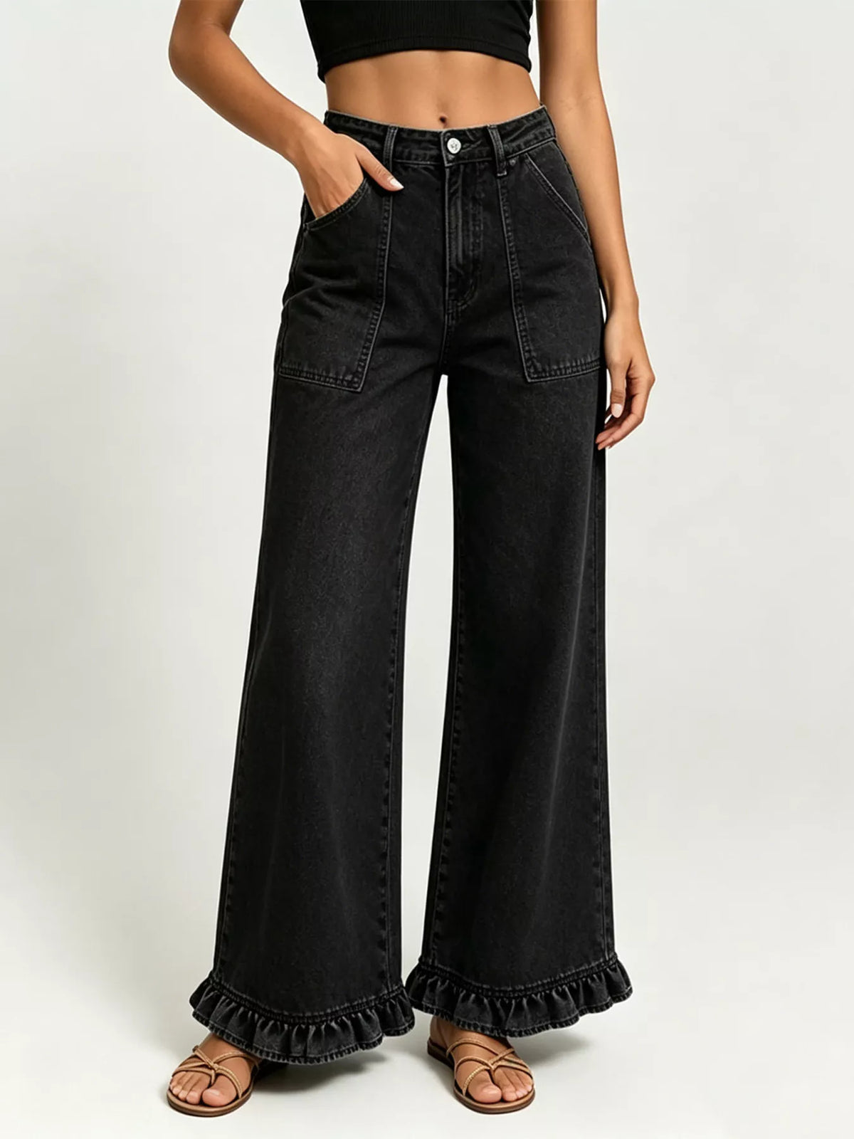 A model wearing black wide-leg denim jeans with a ruffled hem, paired with brown strappy sandals and a black crop top, shown against a light gray studio background.