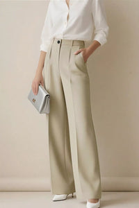 Person wearing a white shirt and beige wide-leg pants holding a silver clutch against a plain background