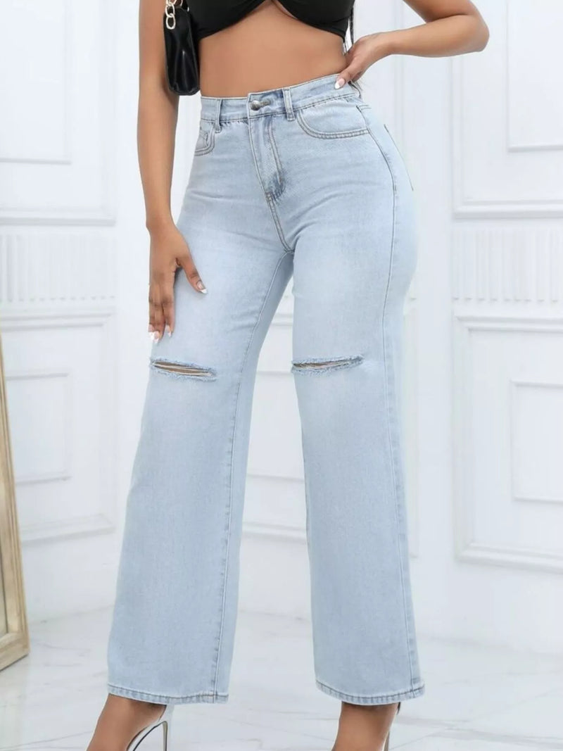 A model wearing high-waisted, wide-leg light blue denim jeans with distressed details on the thighs, paired with a black crop top and white heels.