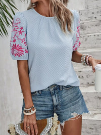 Woman wearing a light blue top with pink floral embroidery and denim shorts, standing on wooden steps.
