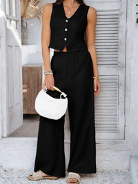 Woman wearing a black sleeveless top and wide-leg pants, holding a white handbag, standing against a light-colored wall.