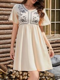Woman wearing a beige dress with floral embroidery standing in front of stacked logs.