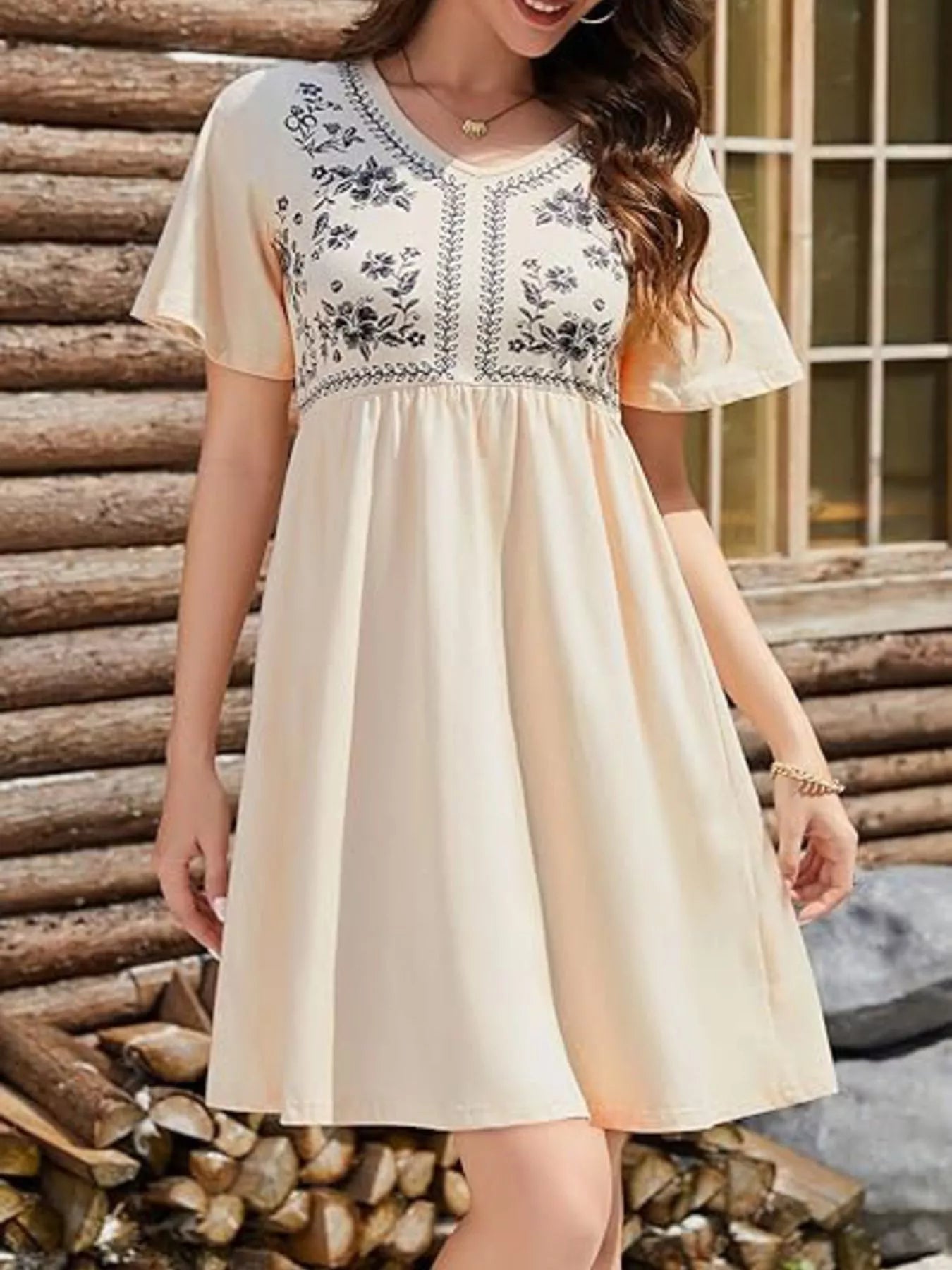 Woman wearing a beige dress with floral embroidery standing in front of stacked logs.