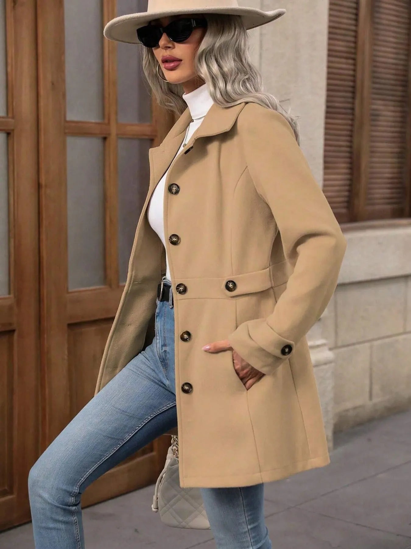 Woman wearing a beige coat, sunglasses, and a hat in an urban setting.