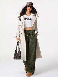 Woman wearing a long beige coat, white crop top, green pants, and brown boots on a white background
