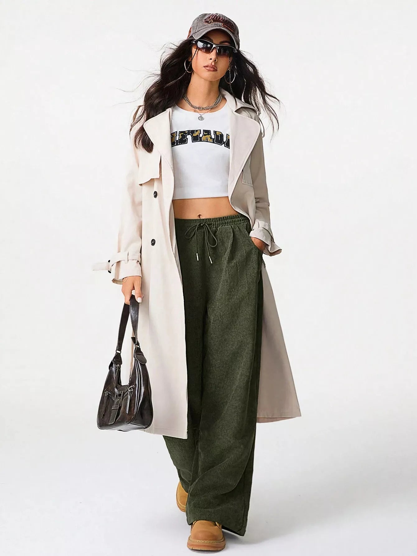 Woman wearing a long beige coat, white crop top, green pants, and brown boots on a white background