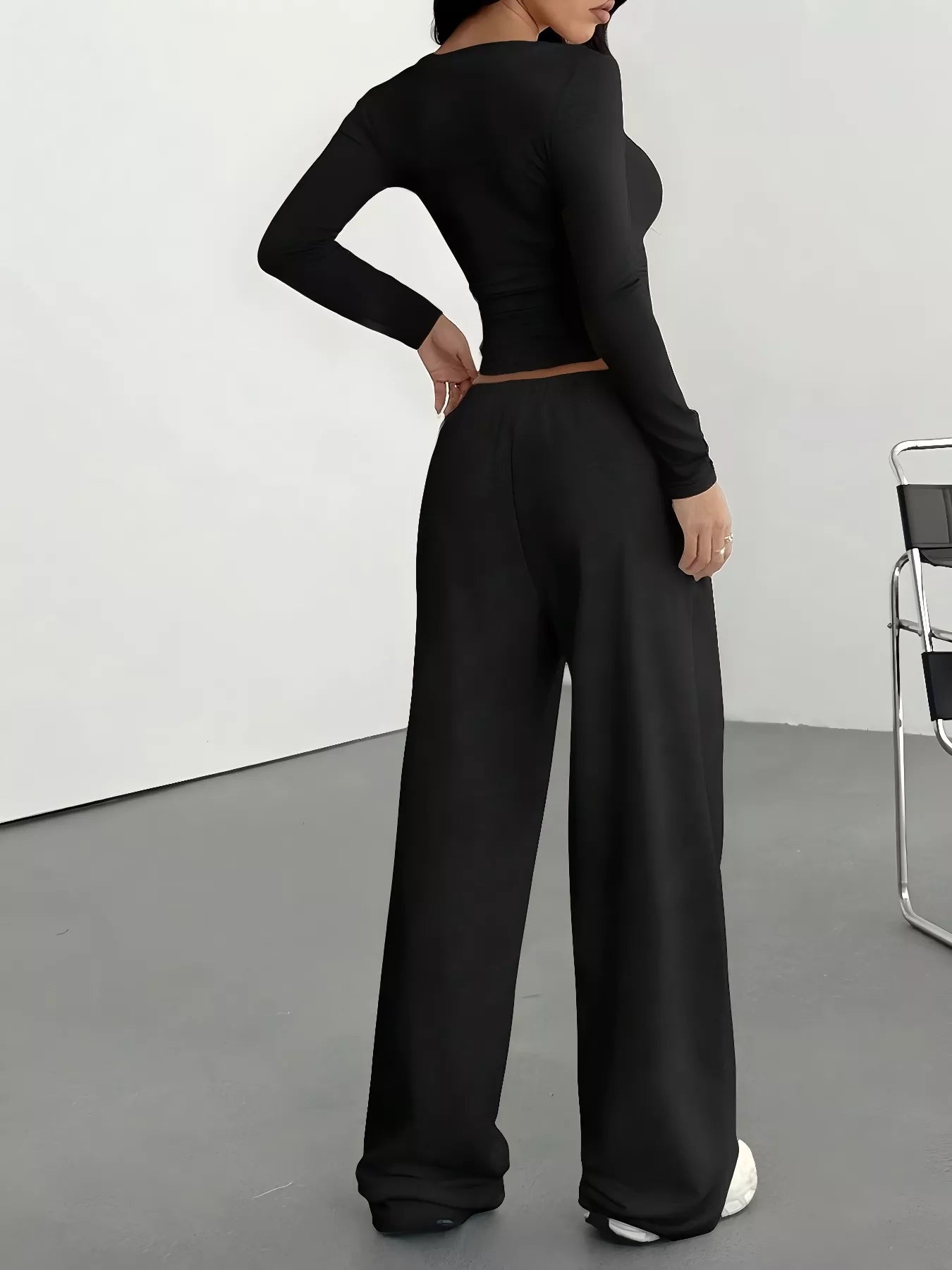 Person wearing a black long-sleeve top and wide-leg pants in a minimalistic setting.