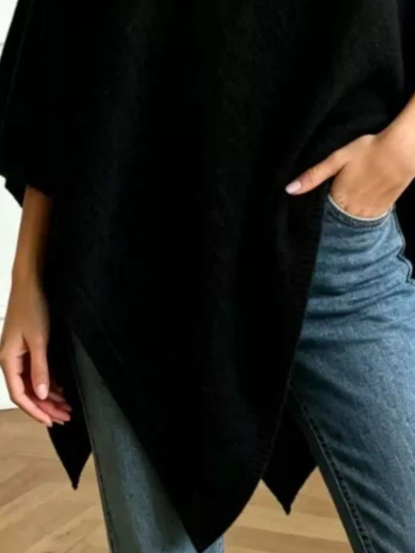 A person wearing a loose black poncho-style top over blue jeans, shown from the chest down with one hand in a pocket.