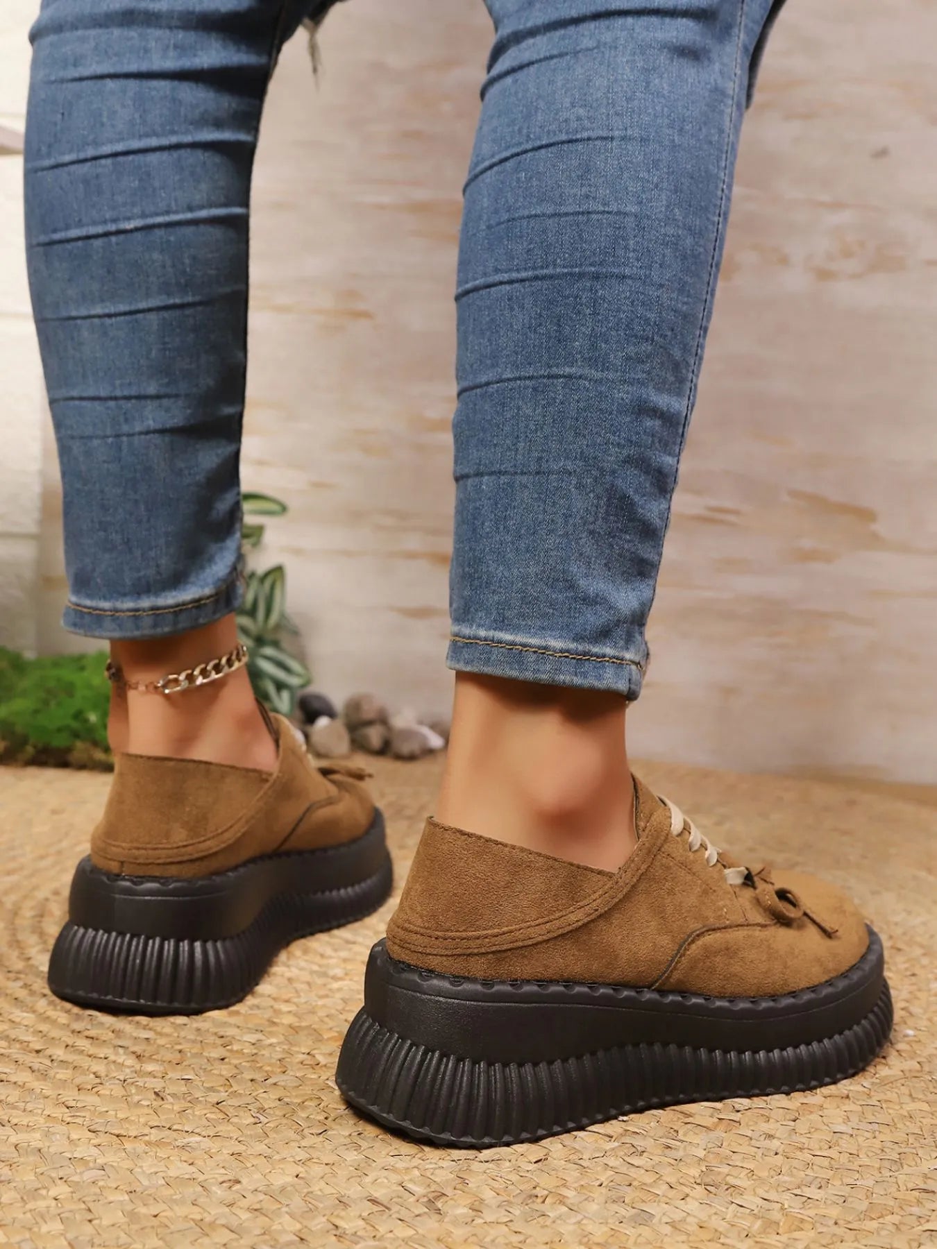 Brown suede shoes with thick black soles worn with blue jeans on a textured surface.