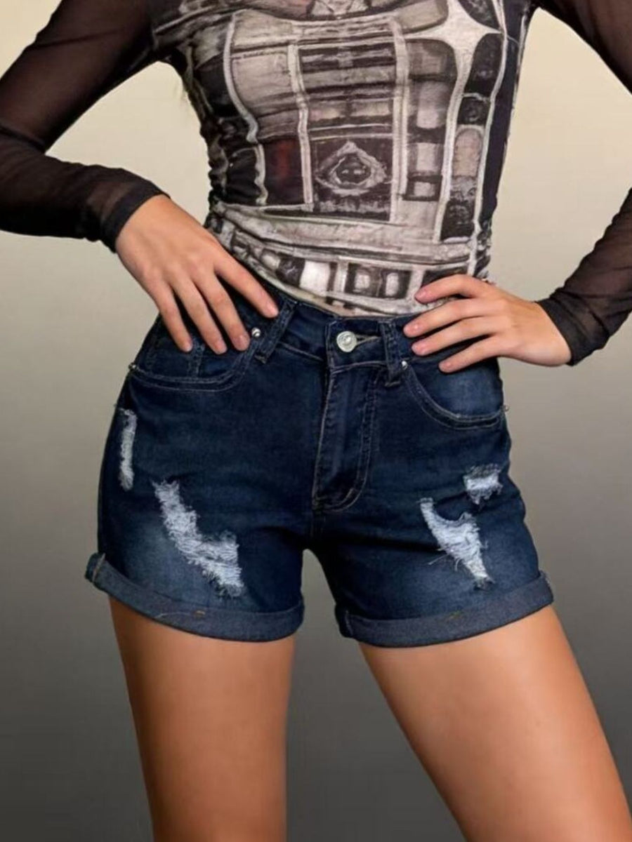 Person wearing a graphic top and distressed denim shorts against a neutral background