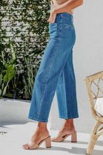 Person wearing blue jeans standing outdoors with plants and a chair in the background