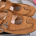 A pair of tan suede Mary Jane-style flats with decorative stitched eye and nose details on the toe, plus buckle straps, shown close up.