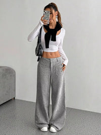 Person wearing a white long-sleeve top with a black bow and gray wide-leg pants in a minimalistic room.