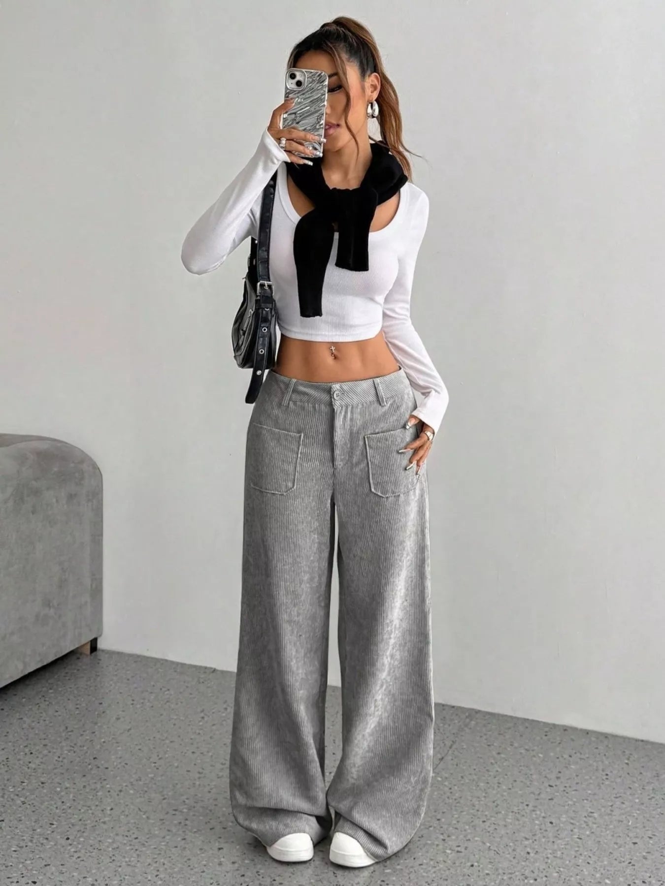 Person wearing a white long-sleeve top with a black bow and gray wide-leg pants in a minimalistic room.