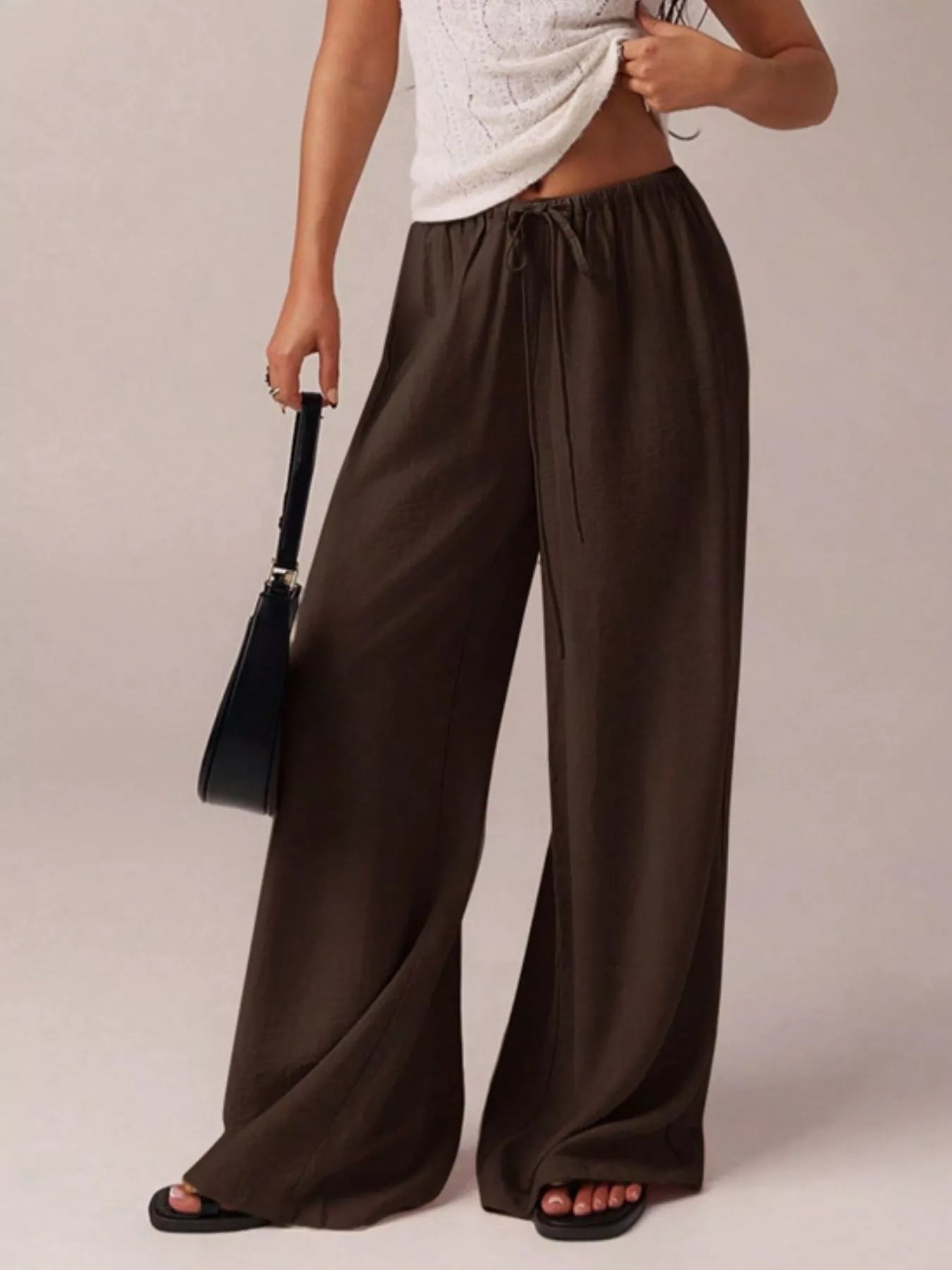 Person wearing brown wide-leg pants holding a black handbag on a plain background