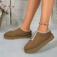 Brown slip-on shoes with fluffy lining worn by a person, with a blurred background of flowers and wooden planks.