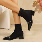 Black pointed-toe ankle boots with a low block heel, shown on a model's feet against a warm beige background with a white chair and pink flowers.