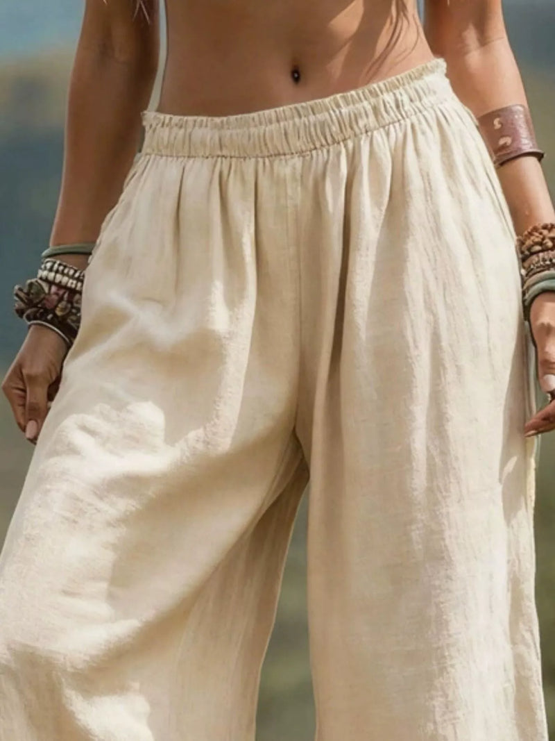 A close-up of a person wearing loose, wide-leg linen pants in a light cream or off-white color. The pants feature an elasticized waistband and a relaxed, draped fit.