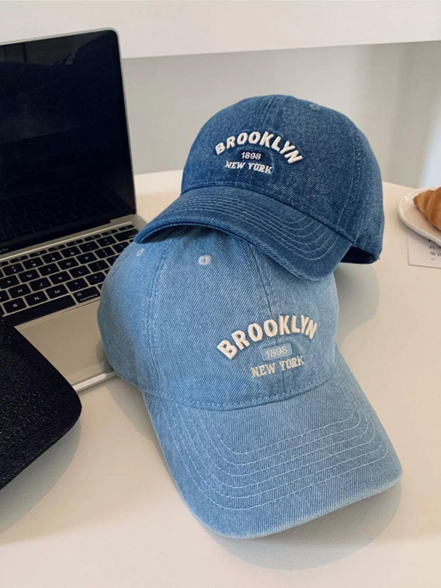 Two blue baseball caps with 'Brooklyn New York' text on a white surface next to a laptop.