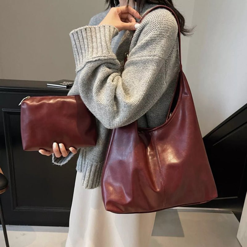 A person wearing a chunky gray knit sweater carries a large, slouchy burgundy leather shoulder bag while holding a matching burgundy leather clutch in one hand.