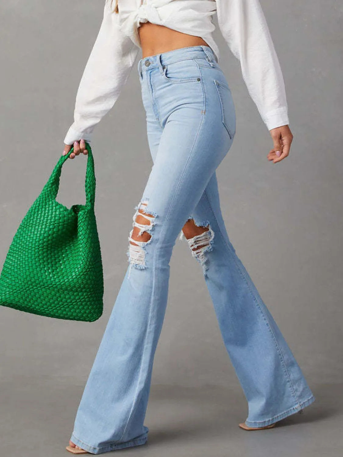 Person wearing light blue flared jeans and holding a green bag on a gray background