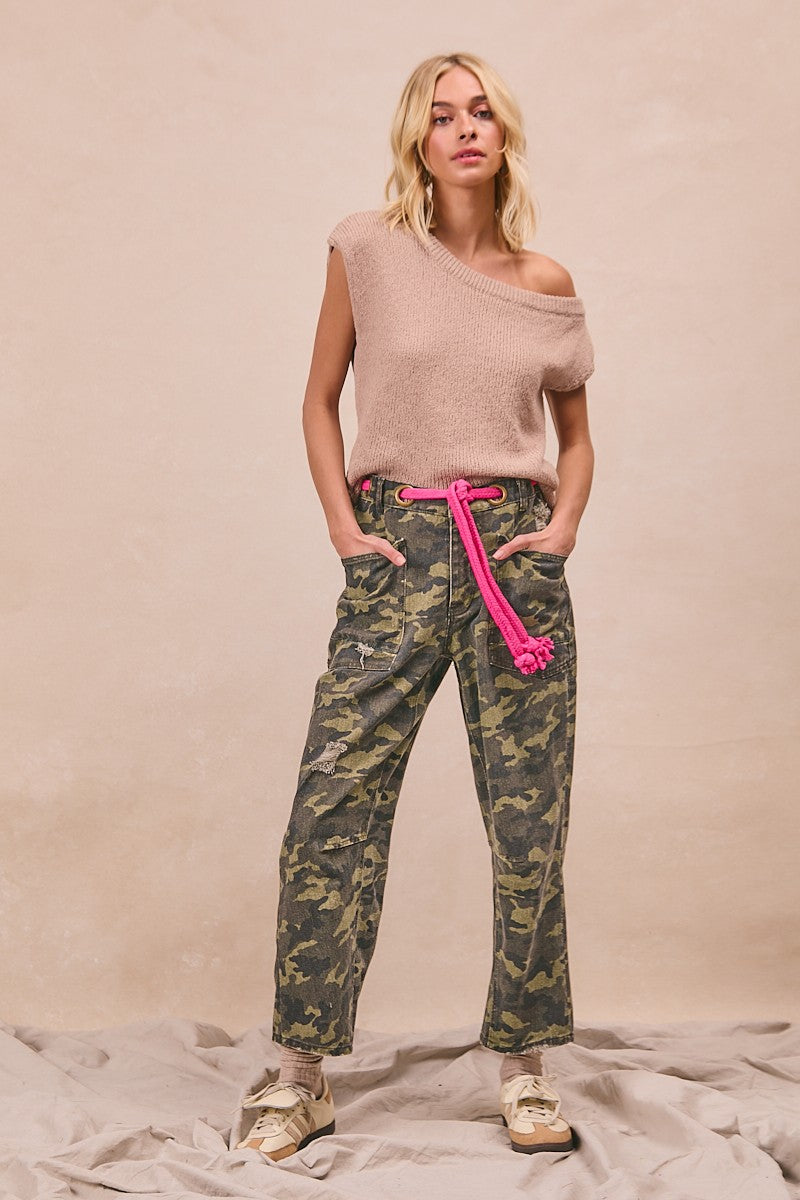 Woman wearing a beige off-shoulder top and camouflage pants with a pink belt against a beige background