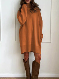 Person wearing a long orange sweater dress with brown knee-high boots against a white wall.