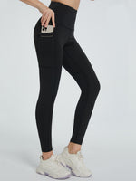A pair of high-waisted black active leggings on a female model, with the model's left hand on her hip. The leggings are designed for sports or active wear, and the model is wearing white sneakers.