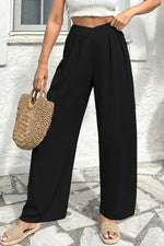 Person wearing black wide-leg pants and a white top, holding a woven bag on a stone pavement.