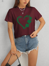 Person wearing a maroon t-shirt with a green heart and shamrock design, paired with denim shorts.