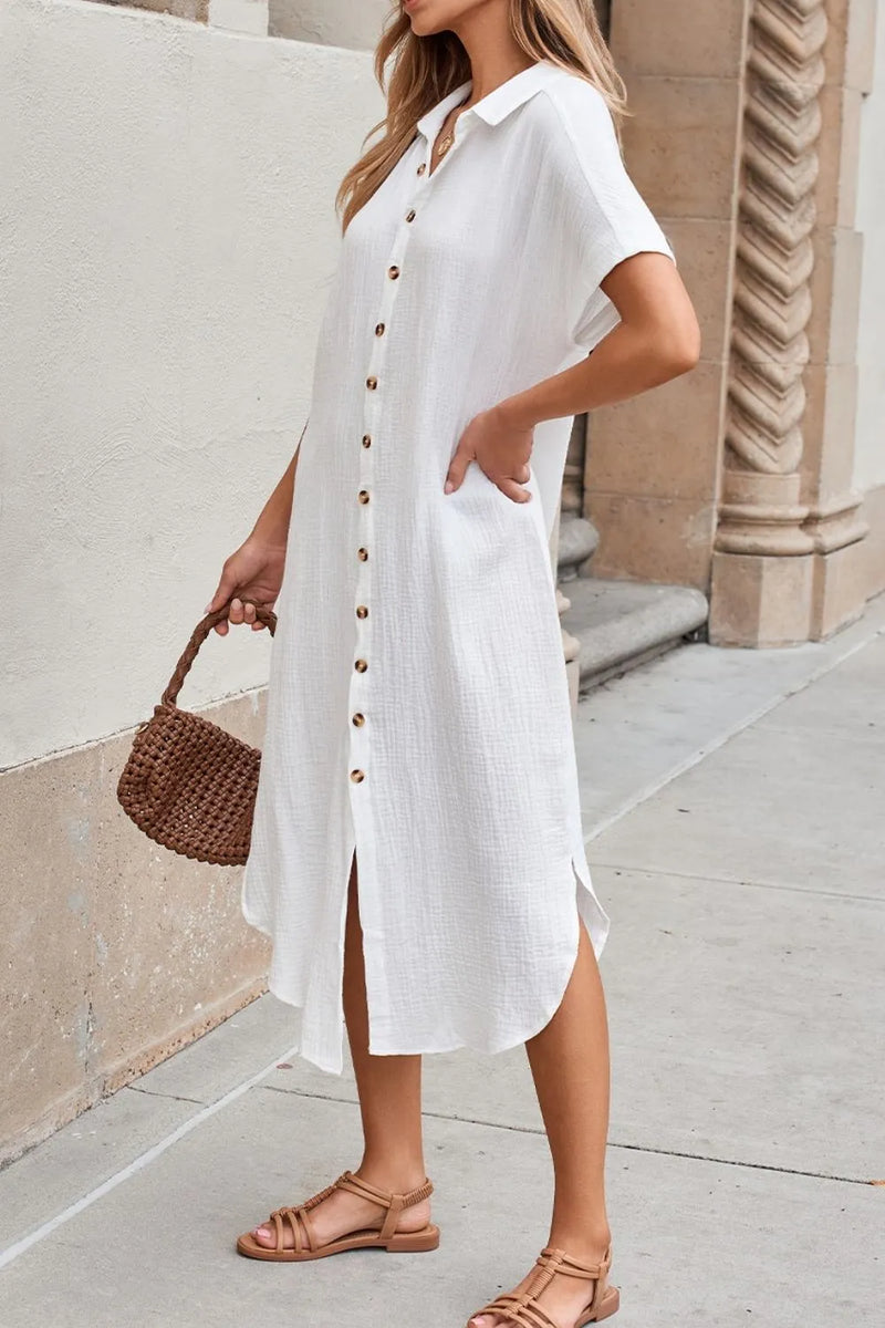 Woman wearing a white dress with buttons, standing against a light-colored wall.