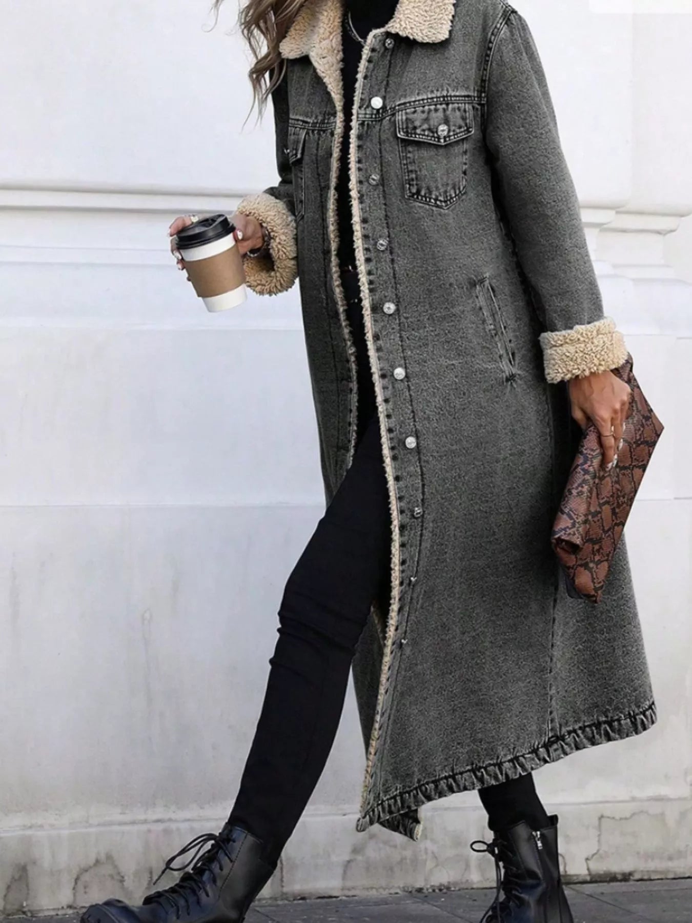 Person wearing a long gray coat with fur trim, holding a coffee cup and clutch.