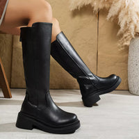 Black knee-high boots worn by a person sitting on a wooden floor.