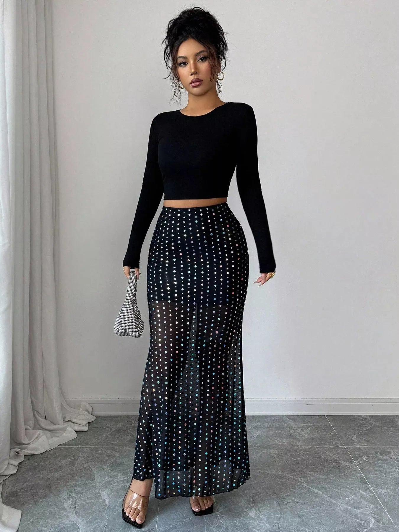 Woman wearing a black long-sleeve top and a black polka dot skirt in a room with light gray curtains.