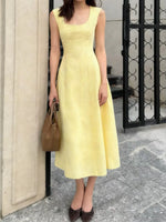 Woman wearing a yellow dress holding a brown handbag against a building exterior.