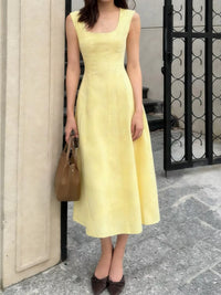 Woman wearing a yellow dress holding a brown handbag against a building exterior.