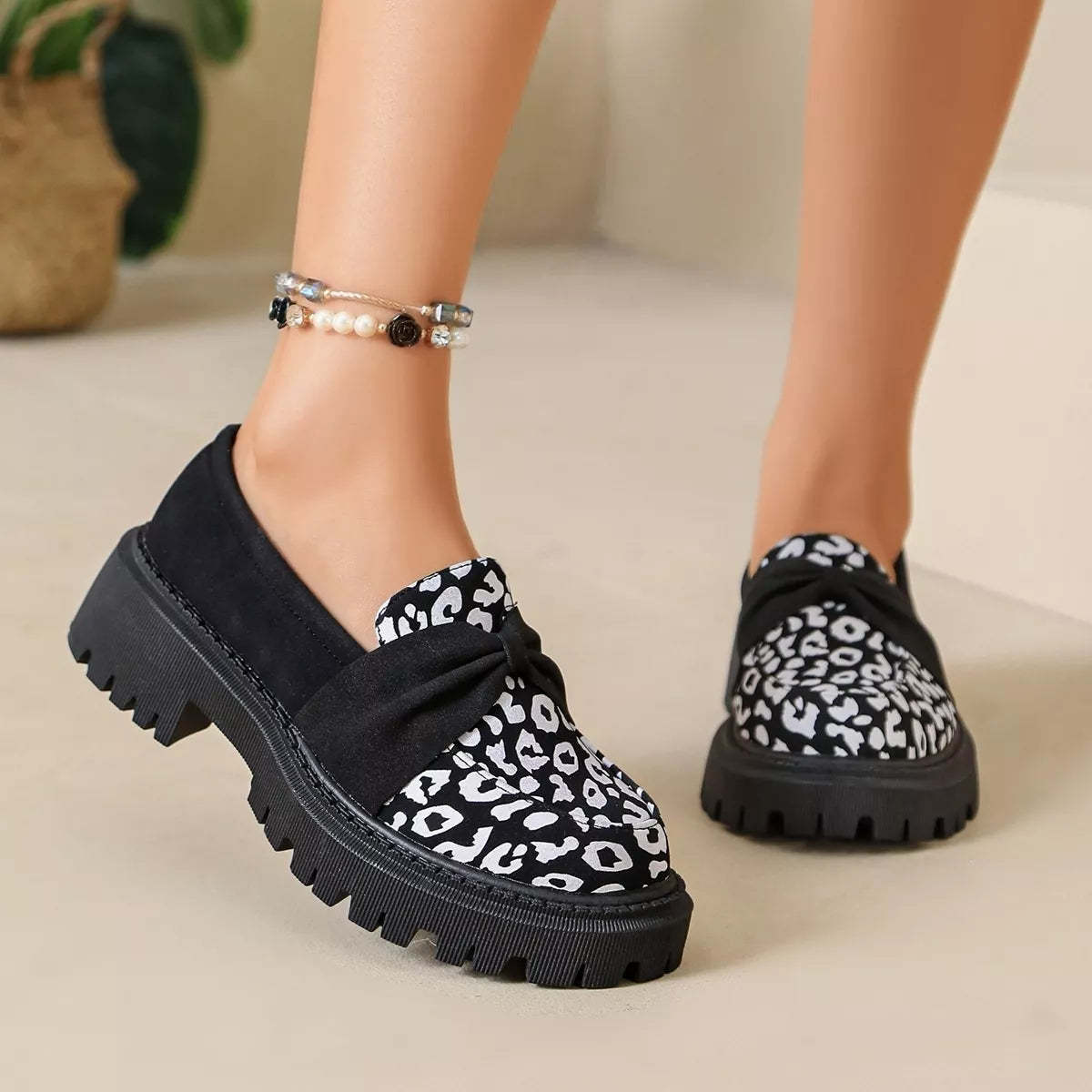 A person wearing black slip-on loafers with a black-and-white leopard print pattern and chunky lug soles, shown from the feet up on a light beige floor.