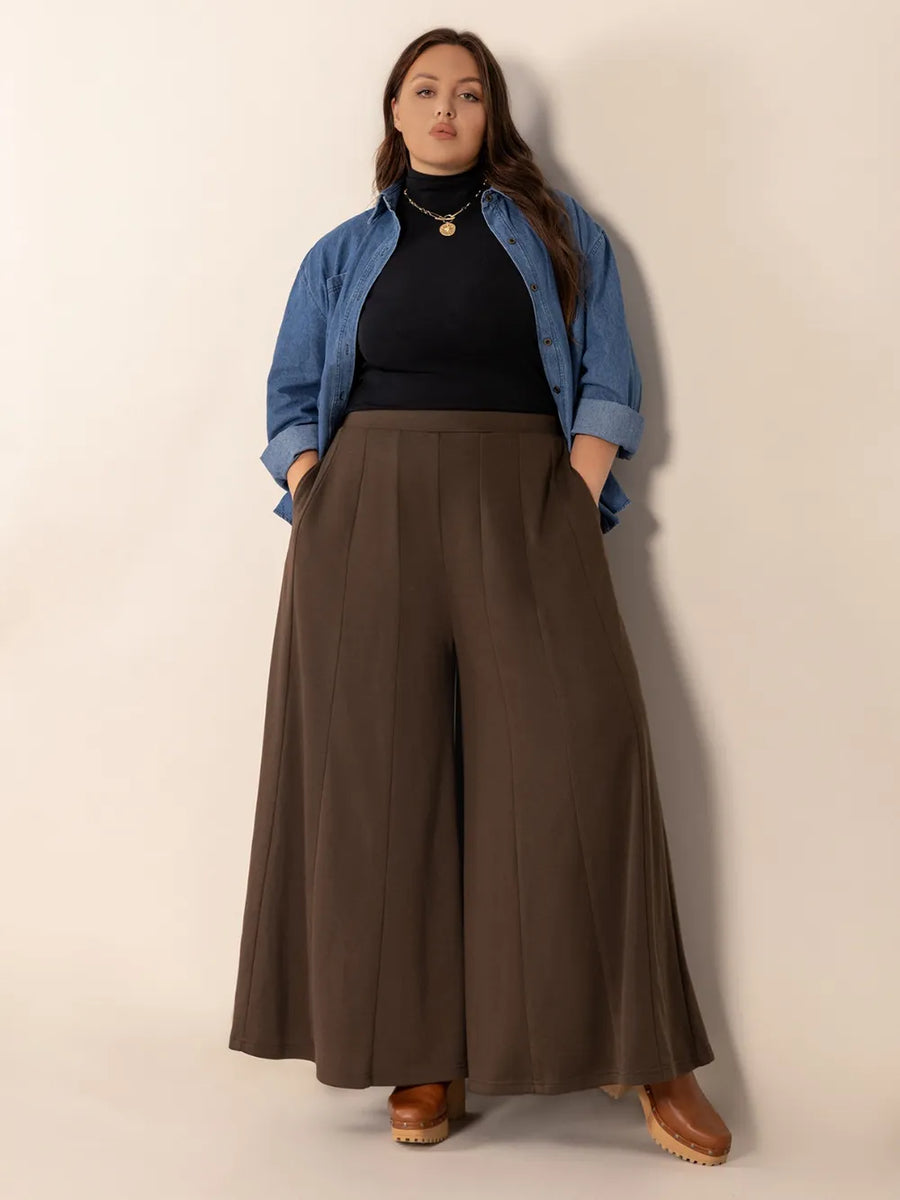 Woman wearing a blue denim jacket, black top, and brown wide-leg pants against a beige background