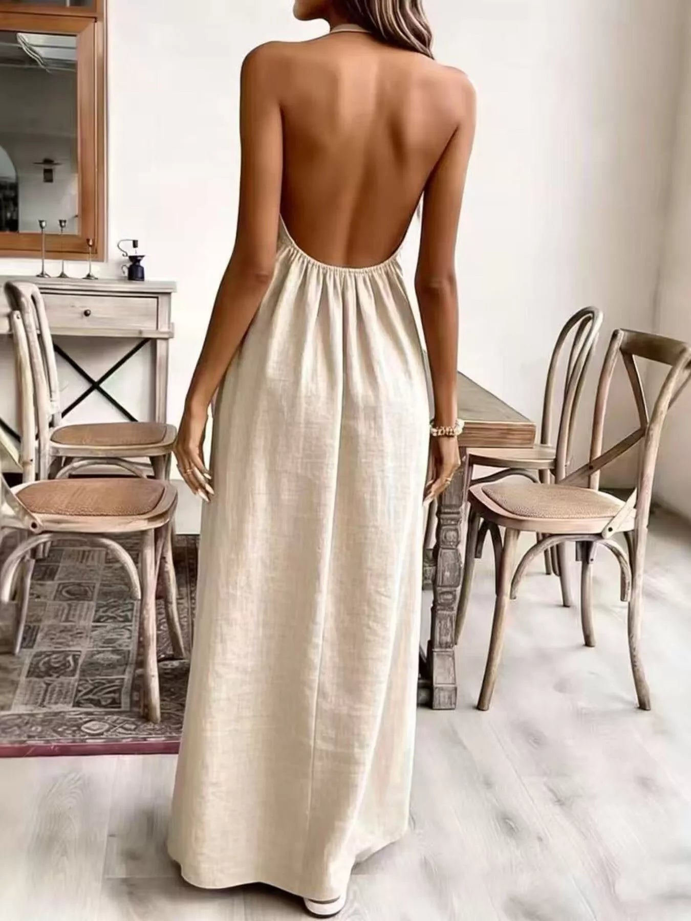 Woman wearing a long, open-back dress in a room with wooden chairs and a mirror.