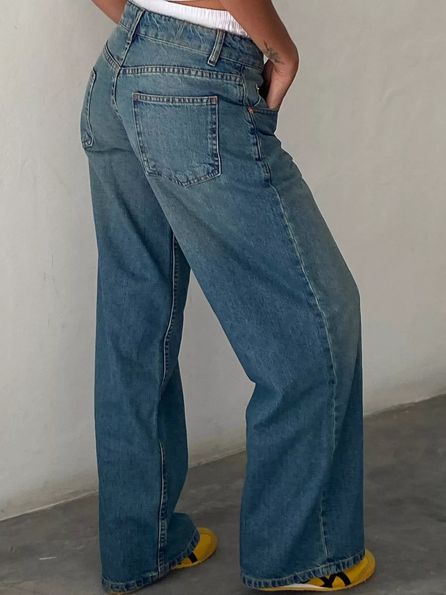 A person wearing blue wide-leg denim jeans, shown from the waist down, standing against a plain off-white wall.